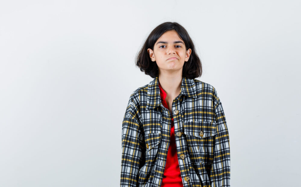young girl standing straight and posing at camera in checked shirt and red t-shirt and looking morose. front view.