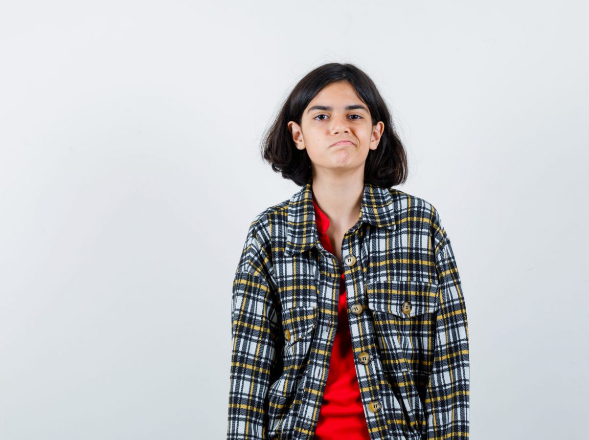 young girl standing straight and posing at camera in checked shirt and red t-shirt and looking morose. front view.